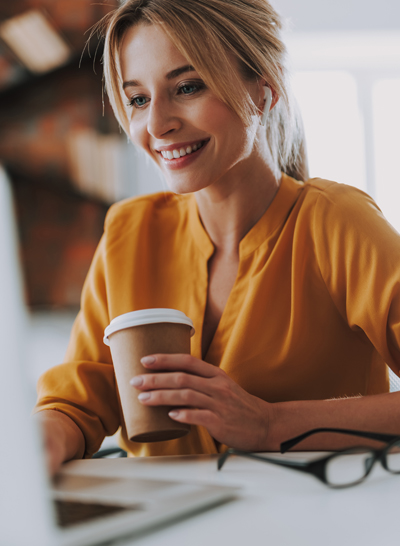 a woman in a yellow shirt holding a coffee cup with her earbuds in smiling at a laptop