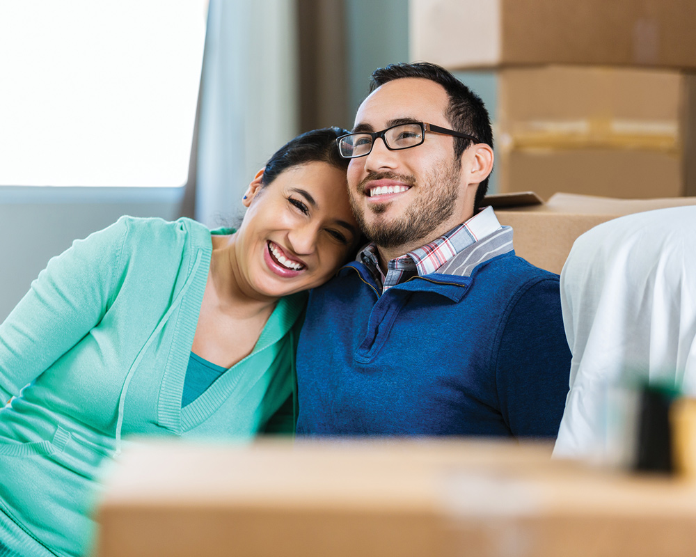 Man in blue with a woman in green resting on his shoulder while they both smile surrounded by moving boxes