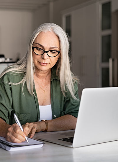 a woman with black glasses and a green shirt writing something down on her notebook in front of her laptop