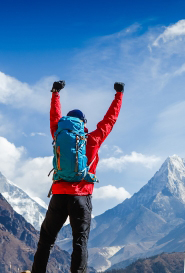 a man in Mountain hiking gear celebrating with his hands raised in the air in front of a mountain skyline.